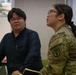 Two individuals are seated in what appears to be a meeting or briefing room, both looking in the same direction. One person is wearing a U.S. Army camouflage uniform (OCP pattern) with a Medical Corps patch on the sleeve, while the other is dressed in civilian clothing. Both wear glasses and appear attentive.