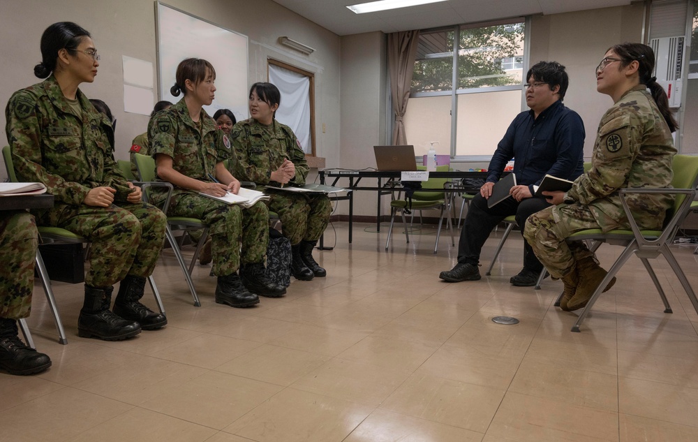 A group of military personnel are seated in a circle in what appears to be a classroom or meeting room, engaged in a discussion or briefing. Three individuals wearing Japanese Ground Self-Defense Force (JGSDF) camouflage uniforms are seated on the left, while one person in US Army MultiCam (OCP) uniform and one civilian are seated on the right. The setting suggests a joint military exchange, training session, or coordination meeting.