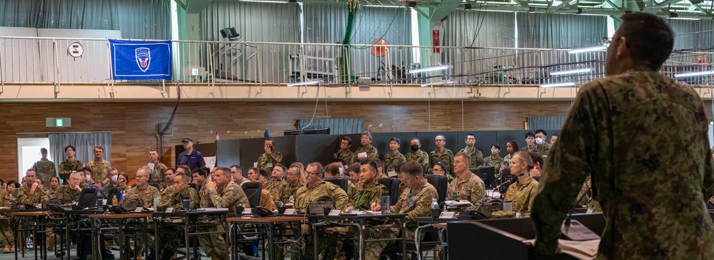 A military briefing or conference is taking place in a large indoor facility, likely a gymnasium or hangar. A speaker stands at a podium addressing a large audience of military personnel in camouflage uniforms seated at tables. The gathering appears to be a multinational military exercise or planning session based on the diverse uniforms visible.