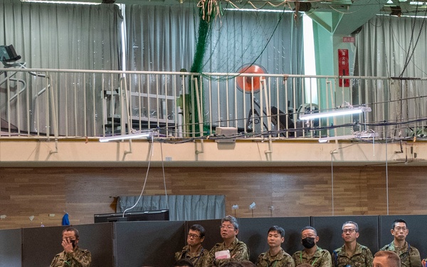 A military briefing or conference is taking place in a large indoor facility, likely a gymnasium or hangar. A speaker stands at a podium addressing a large audience of military personnel in camouflage uniforms seated at tables. The gathering appears to be a multinational military exercise or planning session based on the diverse uniforms visible.