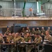 A military briefing or conference is taking place in a large indoor facility, likely a gymnasium or hangar. A speaker stands at a podium addressing a large audience of military personnel in camouflage uniforms seated at tables. The gathering appears to be a multinational military exercise or planning session based on the diverse uniforms visible.