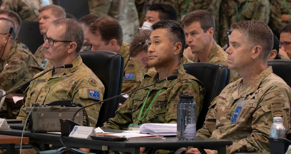 A multinational military briefing or conference is underway, with uniformed personnel from at least two nations seated at a table. Three primary figures are visible in the foreground: two wearing Australian Army multicam uniforms with Australian flag patches, and one wearing a US Army uniform with Ranger, Arctic, and Airborne tabs. A fourth individual in the center appears to be wearing Japanese Ground Self-Defense Force camouflage with a green lanyard badge.