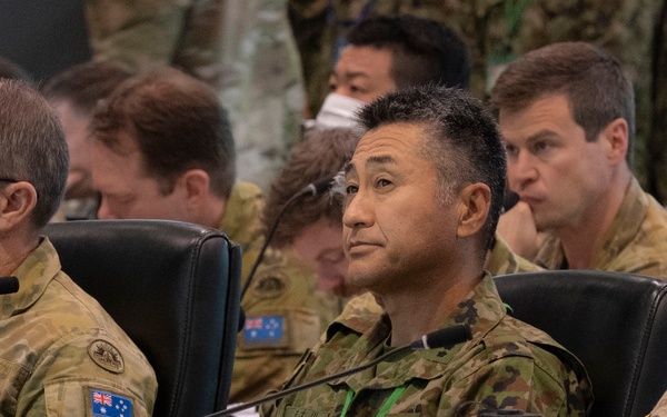 A multinational military briefing or conference is underway, with uniformed personnel from at least two nations seated at a table. Three primary figures are visible in the foreground: two wearing Australian Army multicam uniforms with Australian flag patches, and one wearing a US Army uniform with Ranger, Arctic, and Airborne tabs. A fourth individual in the center appears to be wearing Japanese Ground Self-Defense Force camouflage with a green lanyard badge.