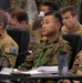 A multinational military briefing or conference is underway, with uniformed personnel from at least two nations seated at a table. Three primary figures are visible in the foreground: two wearing Australian Army multicam uniforms with Australian flag patches, and one wearing a US Army uniform with Ranger, Arctic, and Airborne tabs. A fourth individual in the center appears to be wearing Japanese Ground Self-Defense Force camouflage with a green lanyard badge.