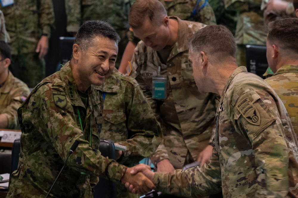 Military personnel from at least two different nations are engaged in what appears to be a multinational military exercise or planning event. A smiling soldier in a distinct camouflage pattern consistent with Japanese Ground Self-Defense Force (JGSDF) uniform is shaking hands with a U.S. Army soldier wearing Ranger, Airborne, and Arctic tabs. The setting appears to be an operations or planning room with multiple uniformed personnel present.