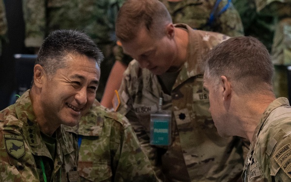 Military personnel from at least two different nations are engaged in what appears to be a multinational military exercise or planning event. A smiling soldier in a distinct camouflage pattern consistent with Japanese Ground Self-Defense Force (JGSDF) uniform is shaking hands with a U.S. Army soldier wearing Ranger, Airborne, and Arctic tabs. The setting appears to be an operations or planning room with multiple uniformed personnel present.