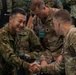Military personnel from at least two different nations are engaged in what appears to be a multinational military exercise or planning event. A smiling soldier in a distinct camouflage pattern consistent with Japanese Ground Self-Defense Force (JGSDF) uniform is shaking hands with a U.S. Army soldier wearing Ranger, Airborne, and Arctic tabs. The setting appears to be an operations or planning room with multiple uniformed personnel present.