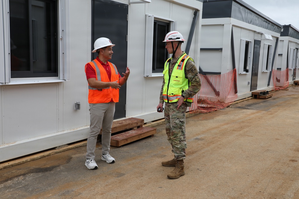 Two individuals are conversing at an active construction or temporary facility site. One person wears civilian clothing with an orange safety vest and white hard hat, while the other is dressed in military camouflage uniform with a yellow high-visibility vest and white hard hat. They appear to be conducting a site inspection or walkthrough alongside a row of modular/prefabricated structures.