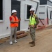 Two individuals are conversing at an active construction or temporary facility site. One person wears civilian clothing with an orange safety vest and white hard hat, while the other is dressed in military camouflage uniform with a yellow high-visibility vest and white hard hat. They appear to be conducting a site inspection or walkthrough alongside a row of modular/prefabricated structures.
