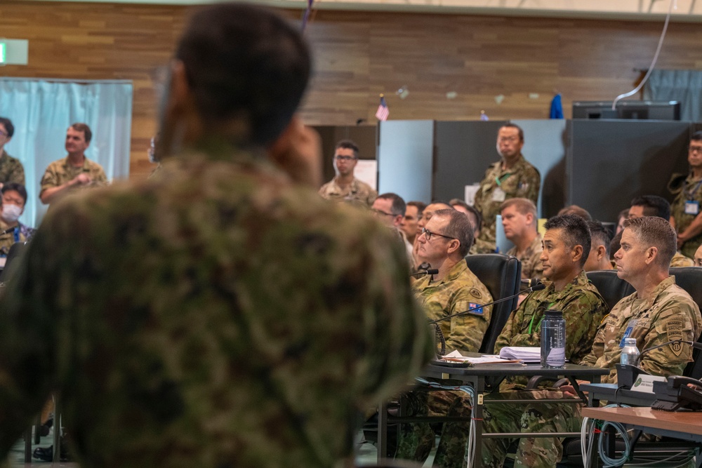 A military briefing or conference is taking place in an indoor facility with personnel from multiple nations present. A speaker in camouflage uniform addresses a seated audience of military personnel, some of whom are seated at tables with documents and microphones. The presence of multiple uniform styles and flag insignia suggests a multinational military exercise or joint operations meeting.