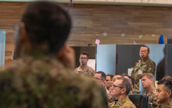 A military briefing or conference is taking place in an indoor facility with personnel from multiple nations present. A speaker in camouflage uniform addresses a seated audience of military personnel, some of whom are seated at tables with documents and microphones. The presence of multiple uniform styles and flag insignia suggests a multinational military exercise or joint operations meeting.