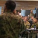 A military briefing or conference is taking place in an indoor facility with personnel from multiple nations present. A speaker in camouflage uniform addresses a seated audience of military personnel, some of whom are seated at tables with documents and microphones. The presence of multiple uniform styles and flag insignia suggests a multinational military exercise or joint operations meeting.