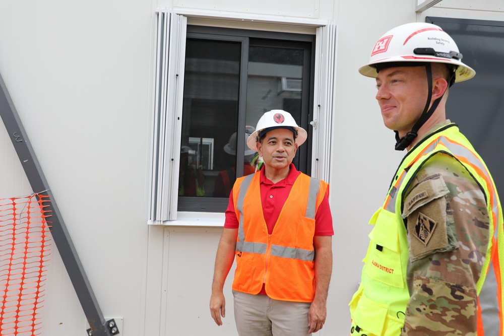Two individuals are standing outside a modular or prefabricated building at what appears to be a construction or military installation site. One person is wearing an orange high-visibility vest and white hard hat, while the other is in U.S. Army camouflage uniform with a yellow high-visibility vest and white hard hat. They appear to be engaged in a site inspection or walkthrough.