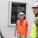 Two individuals are standing outside a modular or prefabricated building at what appears to be a construction or military installation site. One person is wearing an orange high-visibility vest and white hard hat, while the other is in U.S. Army camouflage uniform with a yellow high-visibility vest and white hard hat. They appear to be engaged in a site inspection or walkthrough.