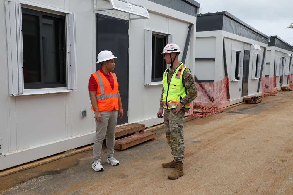 Two individuals are conversing at an active construction or military installation site featuring modular/prefabricated building units. One person is in civilian construction attire with an orange safety vest, while the other is wearing military camouflage (OCP/MultiCam) uniform with a yellow safety vest and white hard hat. The setting appears to be an outdoor compound with temporary or semi-permanent modular structures.