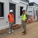 Two individuals are conversing at an active construction or military installation site featuring modular/prefabricated building units. One person is in civilian construction attire with an orange safety vest, while the other is wearing military camouflage (OCP/MultiCam) uniform with a yellow safety vest and white hard hat. The setting appears to be an outdoor compound with temporary or semi-permanent modular structures.