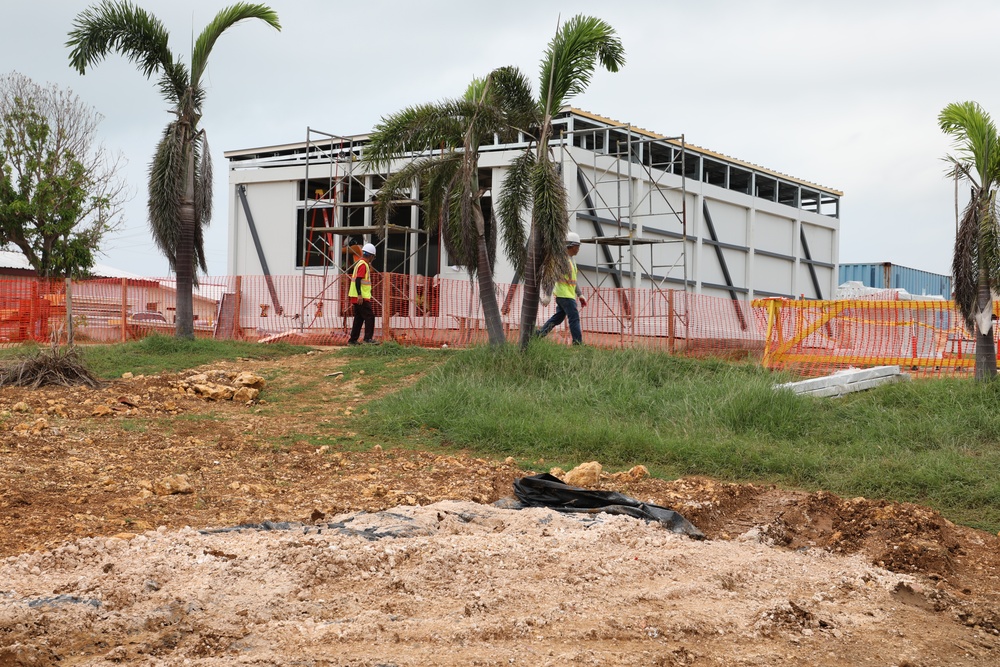 A construction site featuring a partially completed steel-framed building with white panel cladding, surrounded by palm trees in what appears to be a tropical location. Two workers wearing high-visibility vests and hard hats are visible near the structure. The foreground shows disturbed earth, rocky soil, and black plastic sheeting, indicating active ground preparation work.