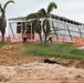 A construction site featuring a partially completed steel-framed building with white panel cladding, surrounded by palm trees in what appears to be a tropical location. Two workers wearing high-visibility vests and hard hats are visible near the structure. The foreground shows disturbed earth, rocky soil, and black plastic sheeting, indicating active ground preparation work.