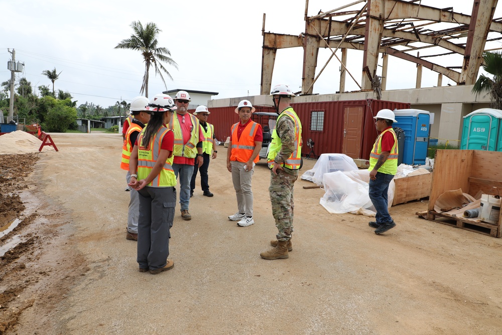 A group of construction and military personnel are gathered at an active construction or disaster recovery site. The scene includes workers in high-visibility safety vests and hard hats, with a damaged steel-frame structure visible in the background. The tropical vegetation and portable toilets suggest this may be a recovery operation in a Pacific island location.
