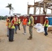 A group of construction and military personnel are gathered at an active construction or disaster recovery site. The scene includes workers in high-visibility safety vests and hard hats, with a damaged steel-frame structure visible in the background. The tropical vegetation and portable toilets suggest this may be a recovery operation in a Pacific island location.