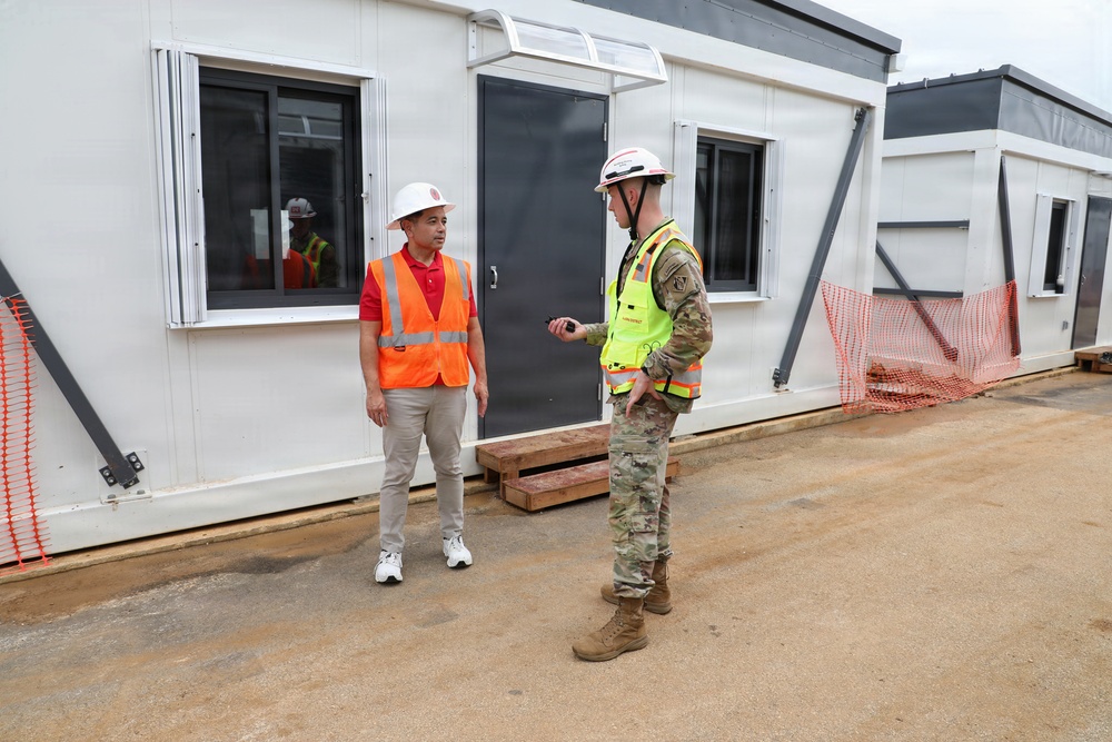 Two individuals are conversing at an active construction or temporary facility site in front of modular/prefabricated buildings. One person is wearing civilian attire with an orange high-visibility vest and white hard hat, while the other is in U.S. Army camouflage uniform with a yellow high-visibility vest and white hard hat. A third person in safety gear is visible in the background through a window.