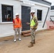 Two individuals are conversing at an active construction or temporary facility site in front of modular/prefabricated buildings. One person is wearing civilian attire with an orange high-visibility vest and white hard hat, while the other is in U.S. Army camouflage uniform with a yellow high-visibility vest and white hard hat. A third person in safety gear is visible in the background through a window.