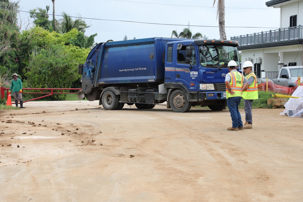 A blue Hyundai garbage/waste collection truck labeled 'ARTMAN' is positioned on a dirt construction or work site. Two workers in high-visibility vests and white hard hats are standing near the truck, apparently overseeing operations. A third worker in green clothing is visible in the background near a red gate barrier.