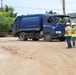 A blue Hyundai garbage/waste collection truck labeled 'ARTMAN' is positioned on a dirt construction or work site. Two workers in high-visibility vests and white hard hats are standing near the truck, apparently overseeing operations. A third worker in green clothing is visible in the background near a red gate barrier.