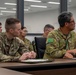 Military personnel from multiple nations are seated at a conference table in a modern briefing room, appearing to attend a joint military meeting or training session. The group includes at least one U.S. Army soldier wearing an 'Old Ironsides' patch and a soldier in Australian Defence Force (ADF) multicam uniform with the name tag 'TOTO'. The setting suggests an international military cooperation or combined arms exercise briefing.