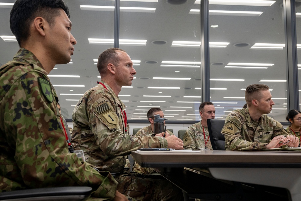 Military personnel from what appears to be the U.S. Army and Japanese Ground Self-Defense Force are seated together in a modern briefing or conference room. The soldiers are attentively facing forward, suggesting they are participating in a joint training session, briefing, or educational event. The setting indicates a formal multinational military cooperation or exchange program.