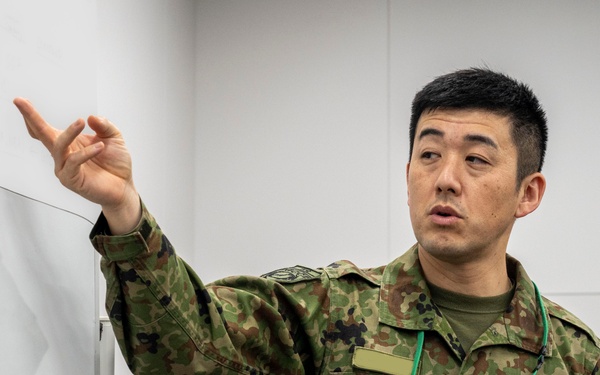A male military personnel wearing Japanese Ground Self-Defense Force (JGSDF) camouflage uniform is gesturing toward a whiteboard or presentation surface. He is wearing an ID badge on a green lanyard and appears to be conducting a briefing or instruction session. The setting appears to be an indoor military or official facility.