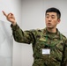 A male military personnel wearing Japanese Ground Self-Defense Force (JGSDF) camouflage uniform is gesturing toward a whiteboard or presentation surface. He is wearing an ID badge on a green lanyard and appears to be conducting a briefing or instruction session. The setting appears to be an indoor military or official facility.