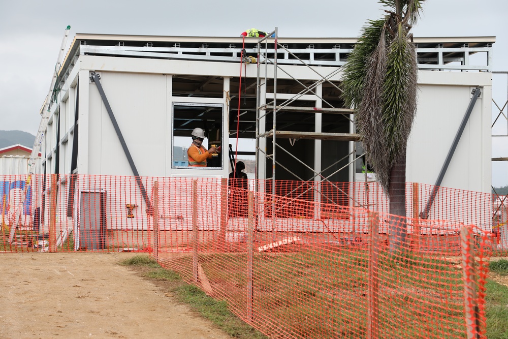 A construction site showing a modular or prefabricated building under assembly. A worker in high-visibility orange clothing and a white hard hat is visible working at window level. The site is secured with orange plastic safety mesh fencing along the perimeter.