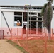 A construction site showing a modular or prefabricated building under assembly. A worker in high-visibility orange clothing and a white hard hat is visible working at window level. The site is secured with orange plastic safety mesh fencing along the perimeter.