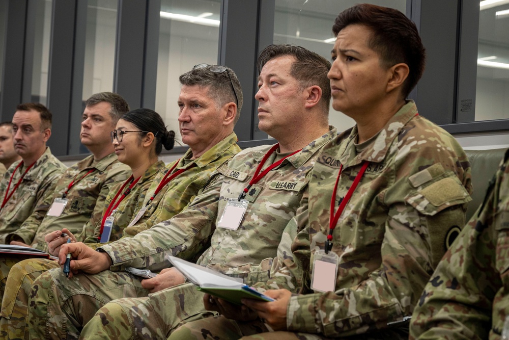 A group of U.S. Army personnel in OCP (Operational Camouflage Pattern) uniforms are seated in rows attending a briefing or conference. Several individuals are wearing red lanyards with name badges and holding notebooks or folders. The setting appears to be a modern indoor facility with large windows.