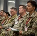 A group of U.S. Army personnel in OCP (Operational Camouflage Pattern) uniforms are seated in rows attending a briefing or conference. Several individuals are wearing red lanyards with name badges and holding notebooks or folders. The setting appears to be a modern indoor facility with large windows.