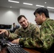 Two military personnel — one U.S. Army soldier and one soldier in a different camouflage uniform — are collaborating at a computer workstation in what appears to be a military operations or command center. The U.S. Army soldier, identified by the name tape 'STREETER' and 'U.S. ARMY' patch, is pointing at a monitor while discussing something with the other soldier. The setting suggests a joint military exercise or coordination activity.
