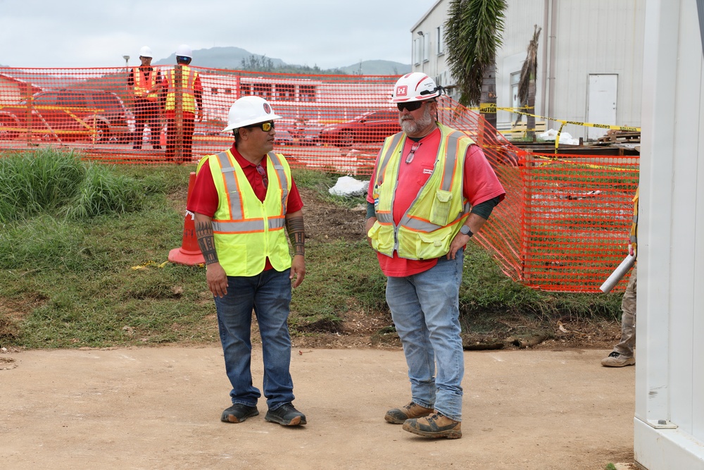 Two construction workers in the foreground are engaged in conversation at an active construction site. Both are wearing standard safety gear including high-visibility yellow vests, hard hats, and red shirts. In the background, a third worker in similar attire is visible near orange safety fencing, with temporary structures and vegetation visible.