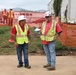 Two construction workers in the foreground are engaged in conversation at an active construction site. Both are wearing standard safety gear including high-visibility yellow vests, hard hats, and red shirts. In the background, a third worker in similar attire is visible near orange safety fencing, with temporary structures and vegetation visible.