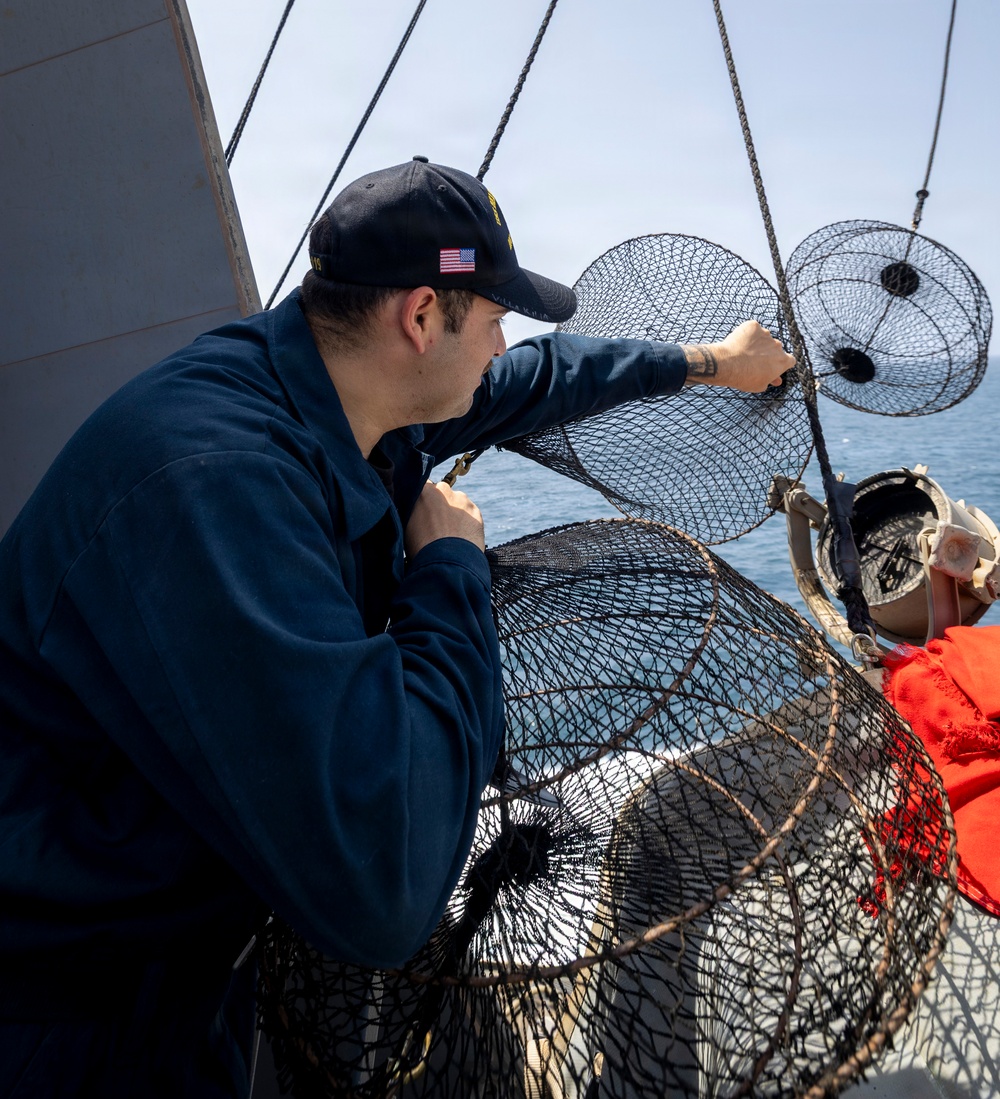 A U.S. Navy sailor wearing a dark navy uniform and a cap with an American flag patch is handling what appear to be crab or lobster traps (wire mesh pots) on the deck of a naval vessel at sea. A second person in an orange garment is partially visible to the right. The scene takes place on open water under a clear sky.
