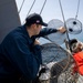 A U.S. Navy sailor wearing a dark navy uniform and a cap with an American flag patch is handling what appear to be crab or lobster traps (wire mesh pots) on the deck of a naval vessel at sea. A second person in an orange garment is partially visible to the right. The scene takes place on open water under a clear sky.