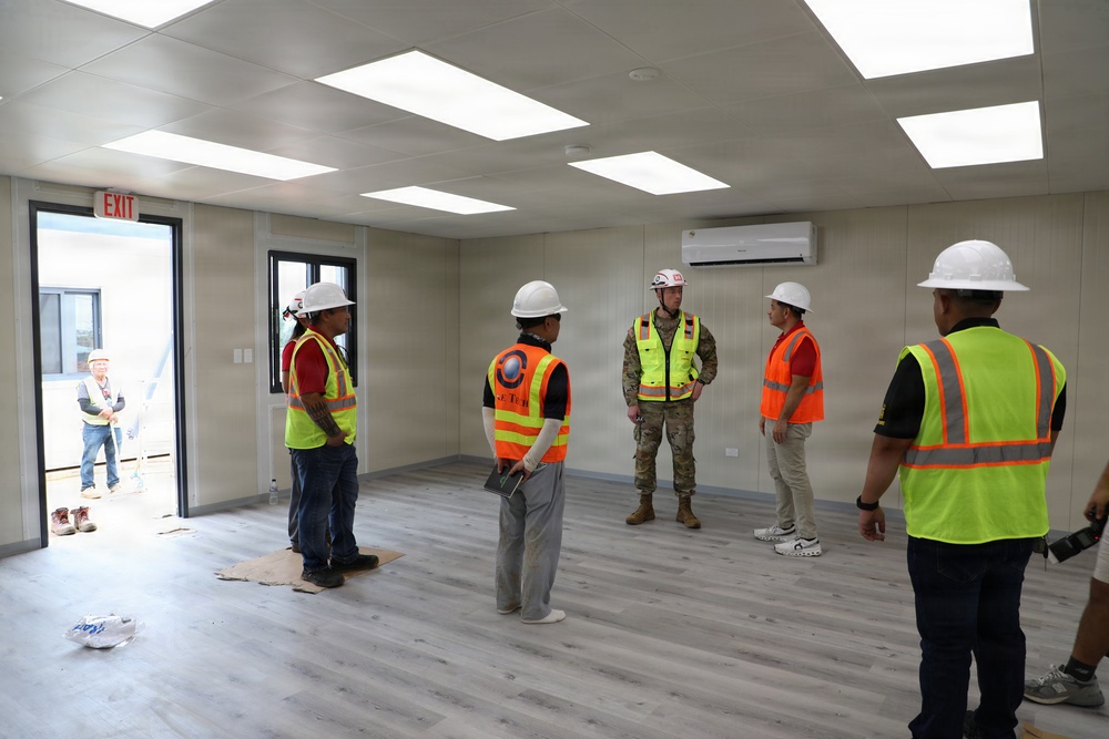 A group of construction workers and a military personnel are gathered inside a newly completed or near-complete modular building for what appears to be an inspection or walkthrough. The interior features finished flooring, ceiling panels with LED lighting, a wall-mounted air conditioning unit, and an EXIT sign near the door. Several individuals are wearing high-visibility safety vests and white hard hats.