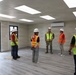 A group of construction workers and a military personnel are gathered inside a newly completed or near-complete modular building for what appears to be an inspection or walkthrough. The interior features finished flooring, ceiling panels with LED lighting, a wall-mounted air conditioning unit, and an EXIT sign near the door. Several individuals are wearing high-visibility safety vests and white hard hats.
