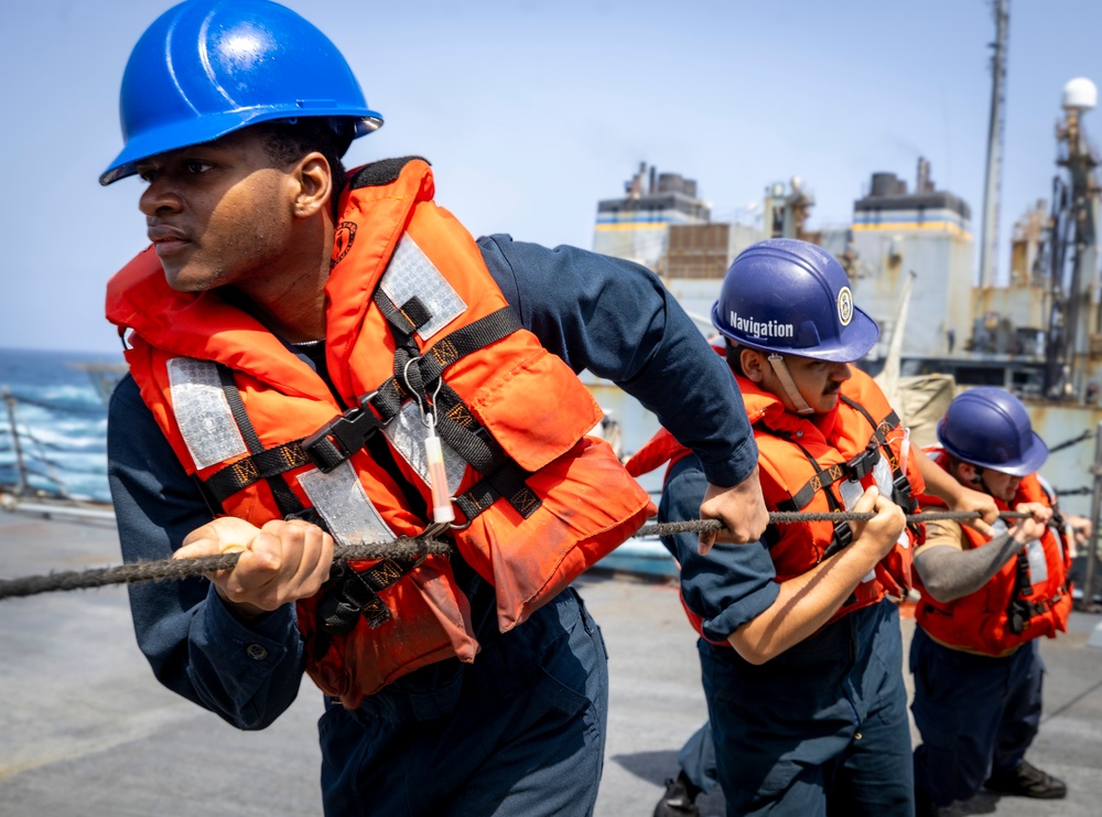 Three naval personnel are pulling a rope in unison on the deck of a ship, wearing orange life vests and hard hats. The foreground sailor wears a blue hard hat, while the middle sailor's helmet is labeled 'Navigation.' A large vessel is visible in the background, suggesting a replenishment-at-sea or mooring operation.