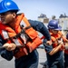 Three naval personnel are pulling a rope in unison on the deck of a ship, wearing orange life vests and hard hats. The foreground sailor wears a blue hard hat, while the middle sailor's helmet is labeled 'Navigation.' A large vessel is visible in the background, suggesting a replenishment-at-sea or mooring operation.