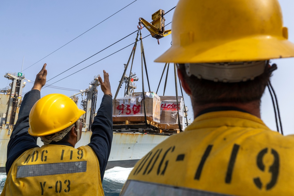 Two personnel wearing yellow hard hats and safety vests marked 'DDG-119' and 'Y-03' are guiding a cargo transfer operation at sea. A large pallet of supplies is being transferred via a highline/replenishment-at-sea (RAS) rigging system between two naval vessels. The operation appears to be a standard underway replenishment (UNREP) evolution.