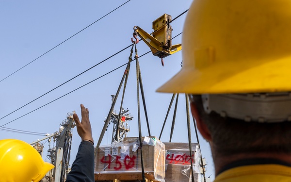 Two personnel wearing yellow hard hats and safety vests marked 'DDG-119' and 'Y-03' are guiding a cargo transfer operation at sea. A large pallet of supplies is being transferred via a highline/replenishment-at-sea (RAS) rigging system between two naval vessels. The operation appears to be a standard underway replenishment (UNREP) evolution.