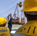 Two personnel wearing yellow hard hats and safety vests marked 'DDG-119' and 'Y-03' are guiding a cargo transfer operation at sea. A large pallet of supplies is being transferred via a highline/replenishment-at-sea (RAS) rigging system between two naval vessels. The operation appears to be a standard underway replenishment (UNREP) evolution.