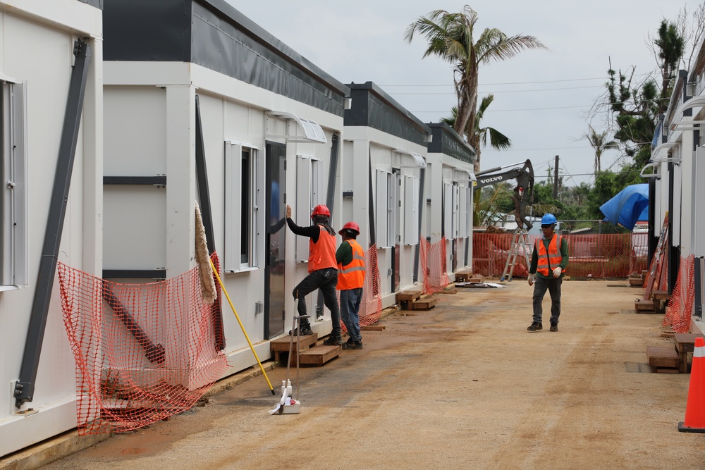Construction workers in high-visibility safety vests and hard hats are installing or finishing modular/prefabricated building units along a dirt pathway. The scene shows an active construction site with multiple white modular structures being assembled side by side. Background elements suggest possible post-disaster reconstruction, including a damaged palm tree and a blue tarp visible in the distance.