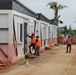 Construction workers in high-visibility safety vests and hard hats are installing or finishing modular/prefabricated building units along a dirt pathway. The scene shows an active construction site with multiple white modular structures being assembled side by side. Background elements suggest possible post-disaster reconstruction, including a damaged palm tree and a blue tarp visible in the distance.